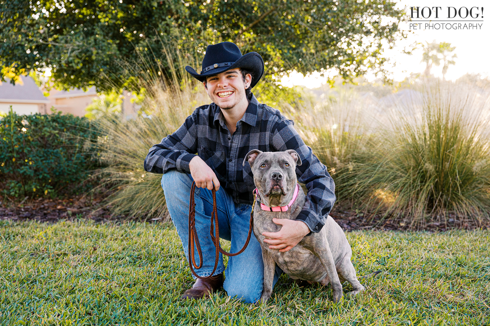 Young man kneels on the grass beside a gray pitbull wearing a pink collar during an outdoor pet photo session.
