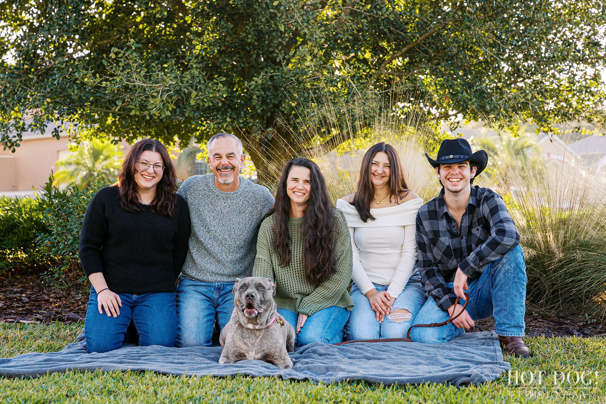 Family sits together on a blanket with their gray pitbull centered in front during an outdoor portrait session.