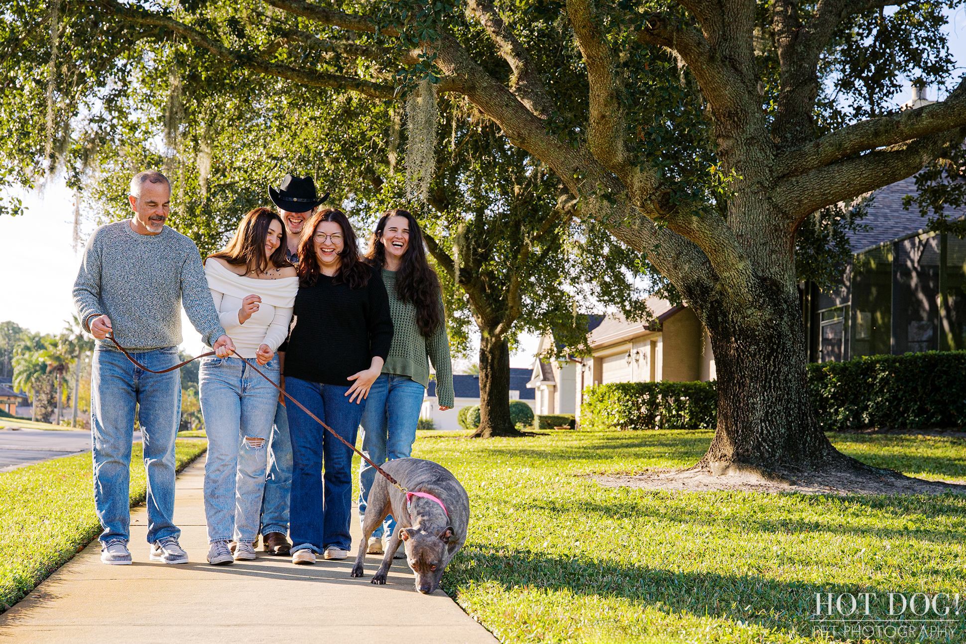 Family walks together along a sidewalk with their gray pitbull on leash during a casual neighborhood photo session.