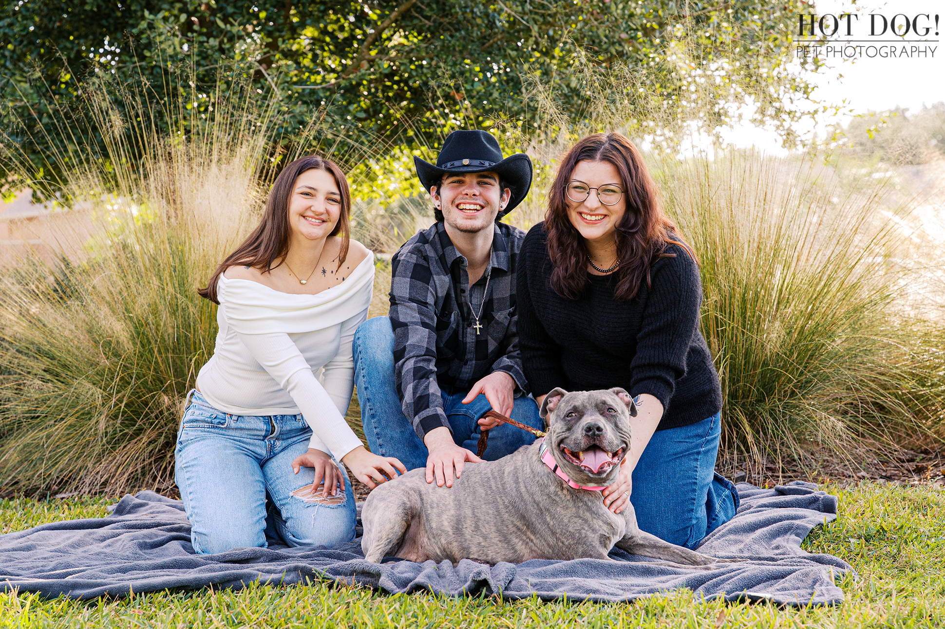 Three young adults sit on a blanket with a gray pitbull lying happily in front of them during a relaxed outdoor portrait.