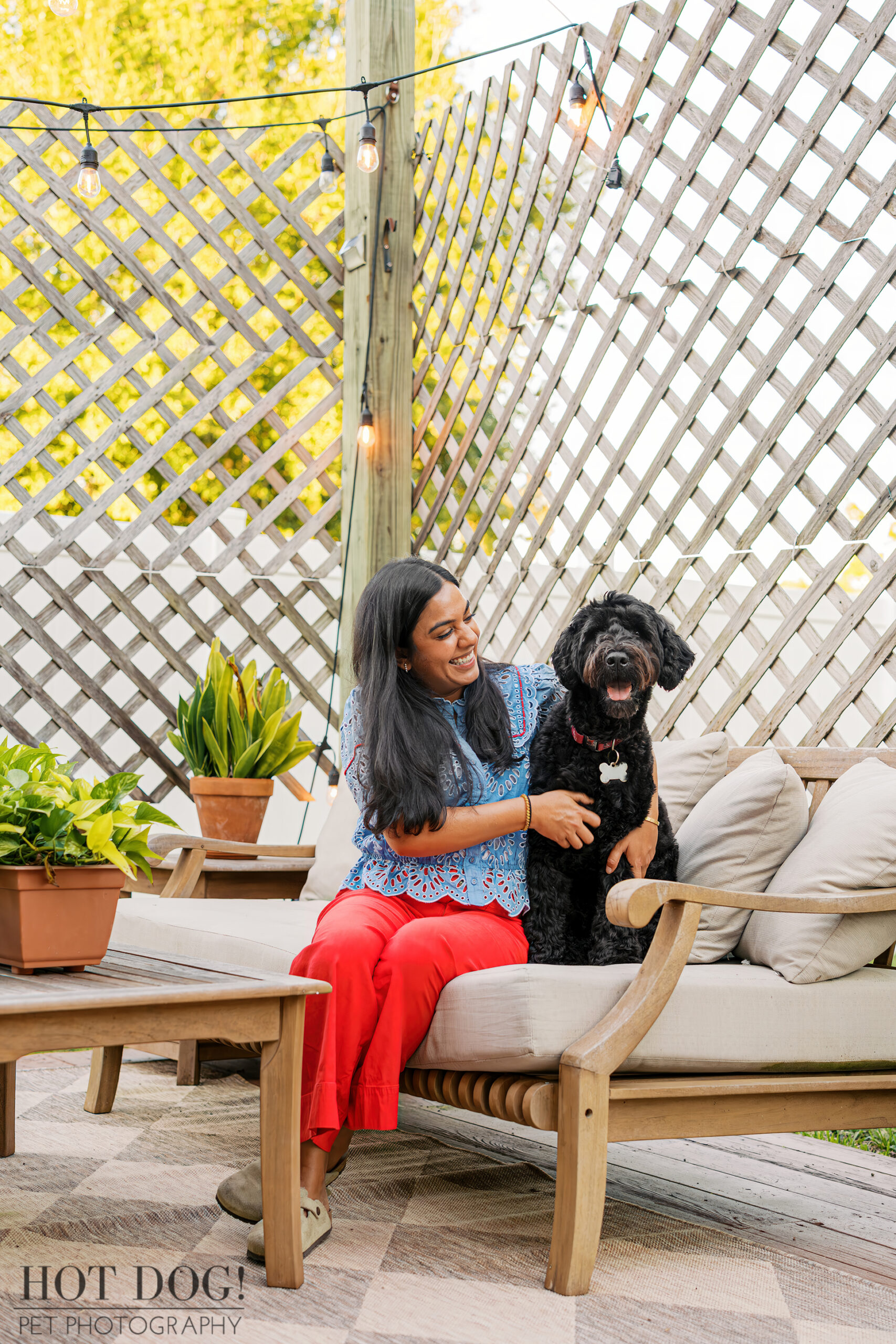 Selavi and Banjo sit together on the outdoor sofa under string lights, sharing a joyful moment surrounded by plants.