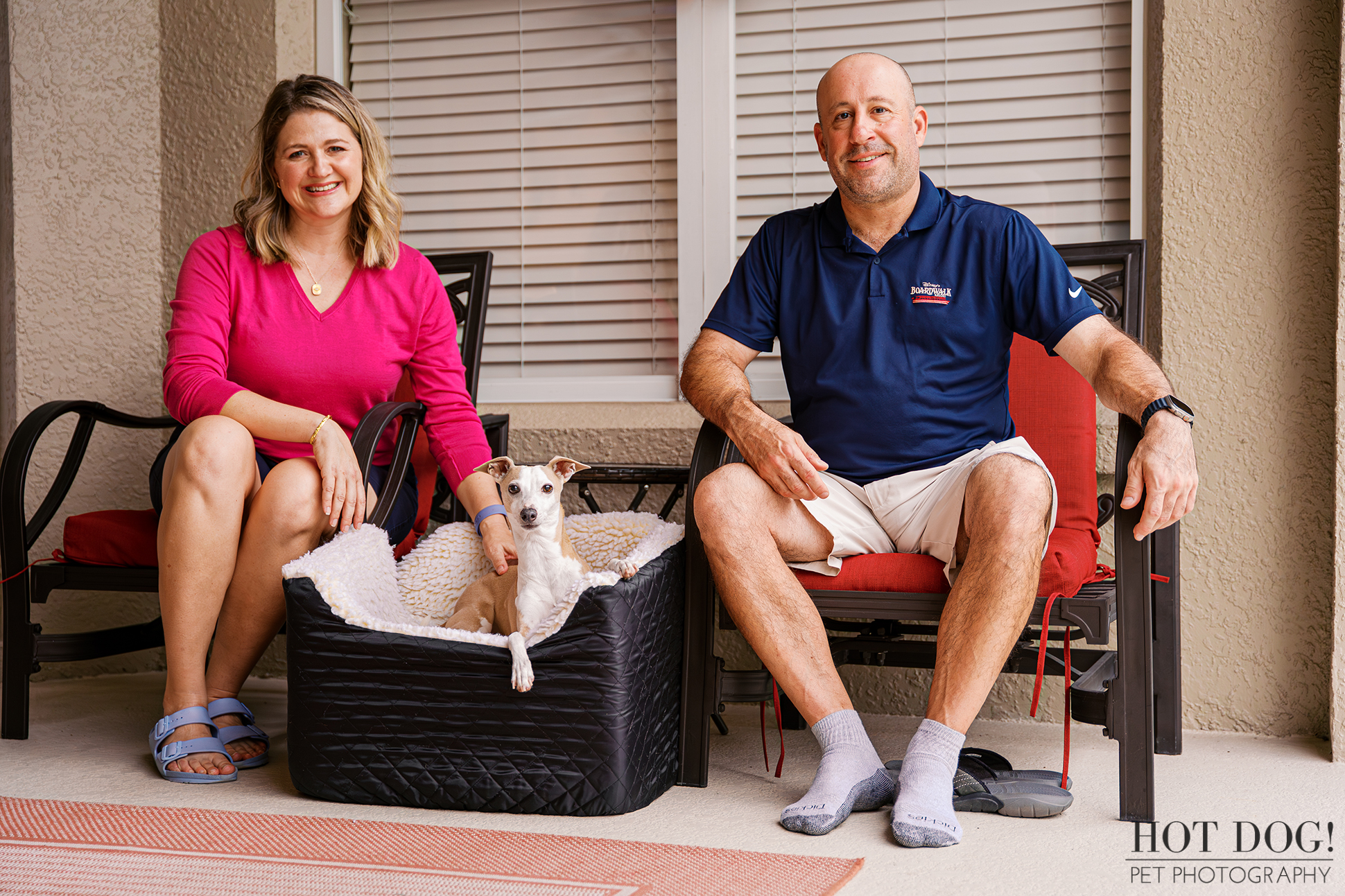 Italian Greyhound being held by his owners in front of a red front door with holiday decor during a home pet photography session.
