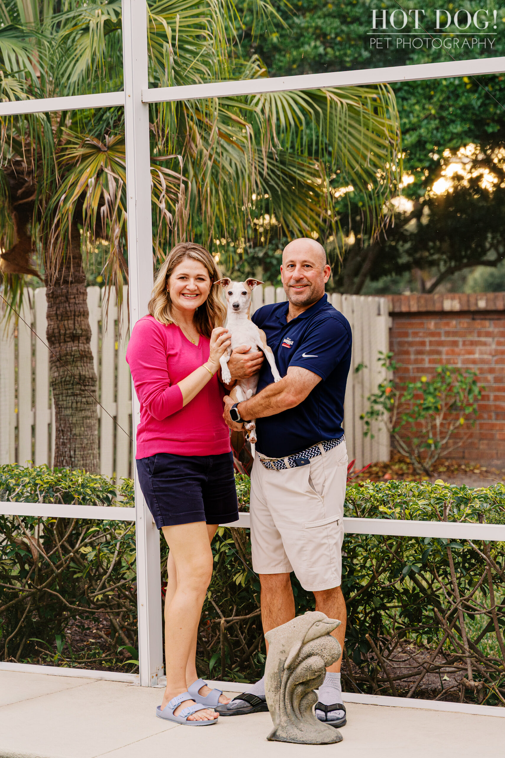 Italian Greyhound being held by his owners in a screened patio area, posing together during an at-home pet photography session.