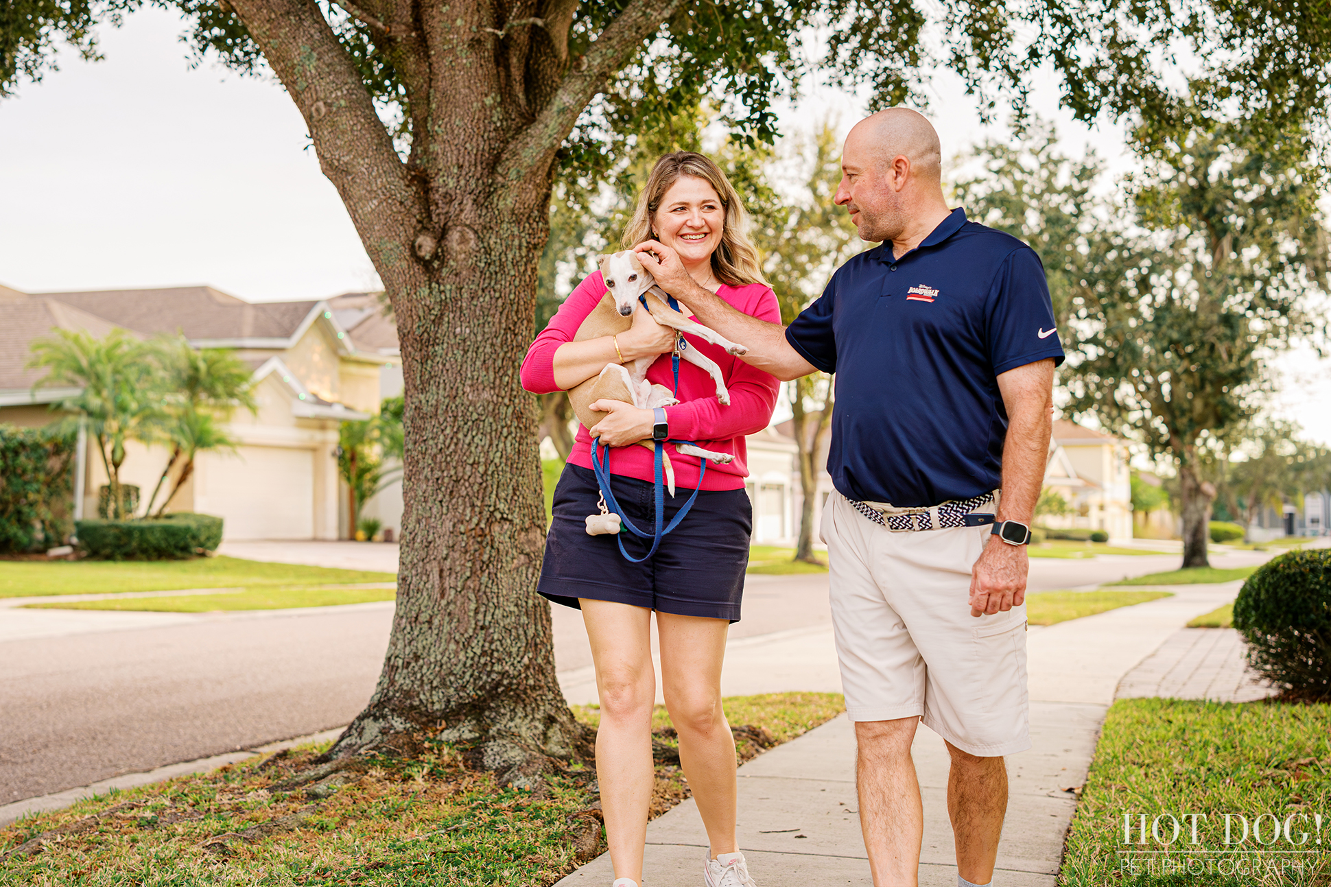 Couple walking along a neighborhood sidewalk while one owner carries their Italian Greyhound during a lifestyle pet photography session