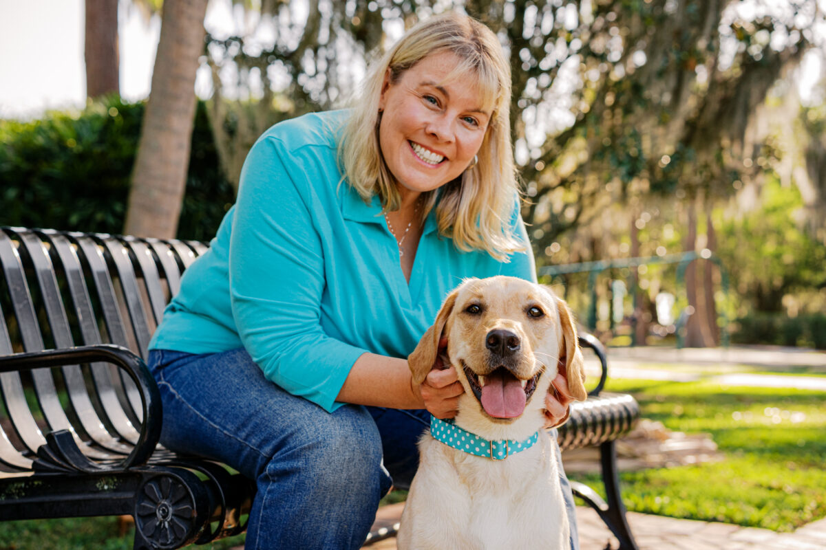 Smiling woman posing beside her happy yellow Labrador Retriever on a park bench during an outdoor pet photography session.
