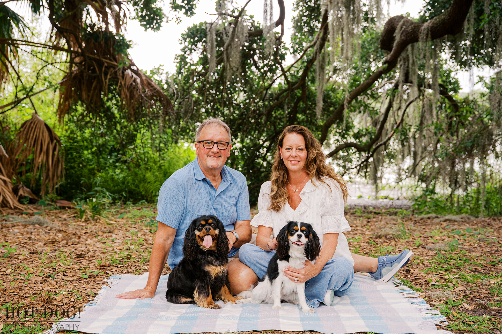 Tonia and James sitting with Cavalier King Charles Spaniels Jazmyn and Jerzee on a picnic blanket beneath Spanish moss in Central Florida.