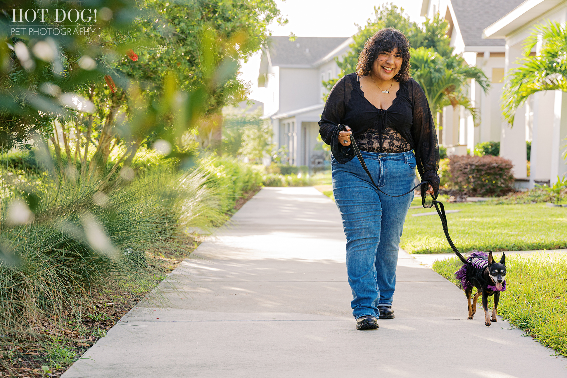 Gabriela walking Kiki down a sunny, tree-lined sidewalk, with Kiki wearing a purple harness and black tutu.