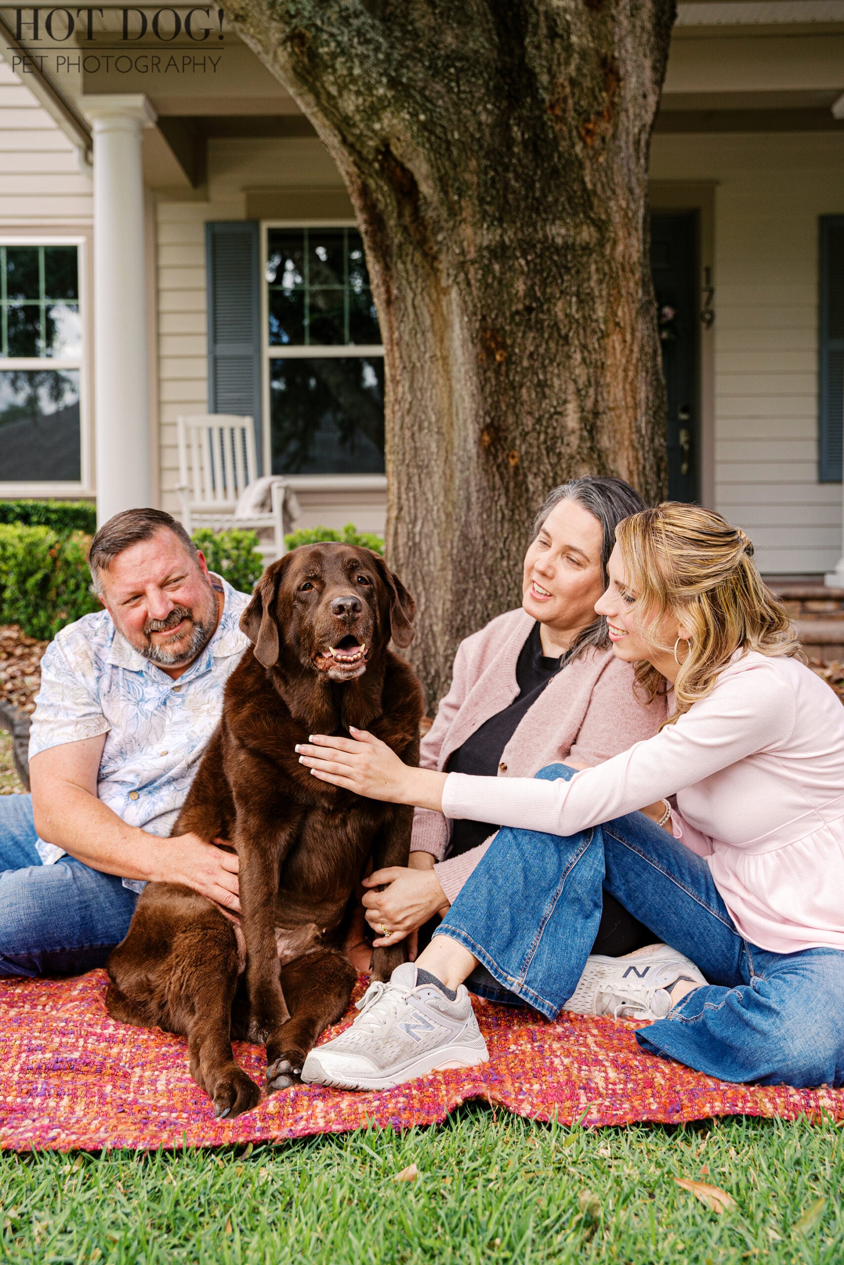 Senior chocolate Labrador Retriever Kona sits beneath a large tree with his loving family during a relaxed outdoor portrait session at their Oviedo home.