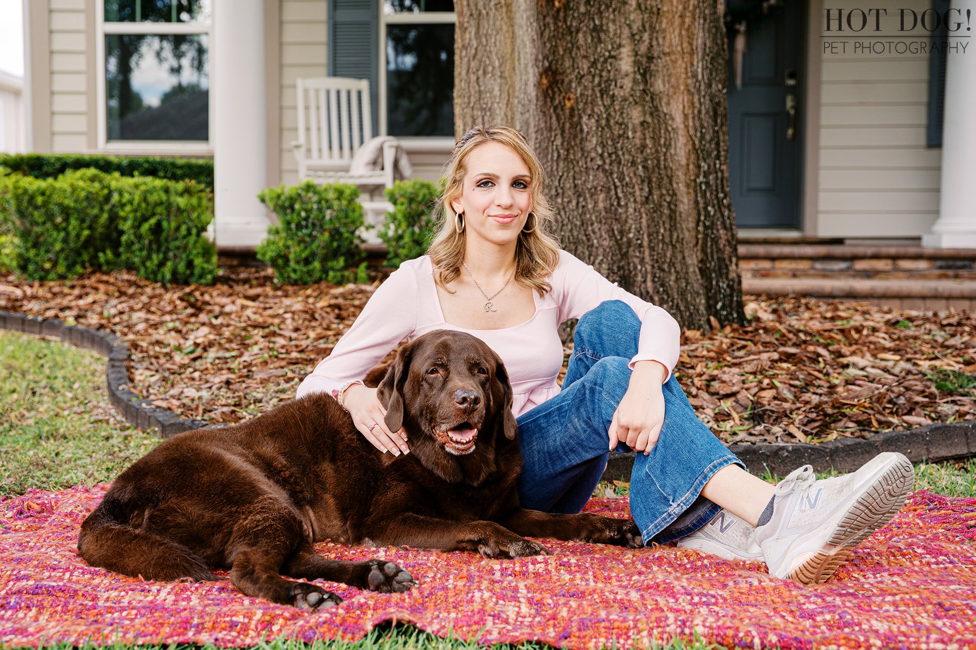 Regina sits beside her senior chocolate Labrador Retriever, Kona, on a colorful blanket beneath a large tree during a family pet portrait session at home.