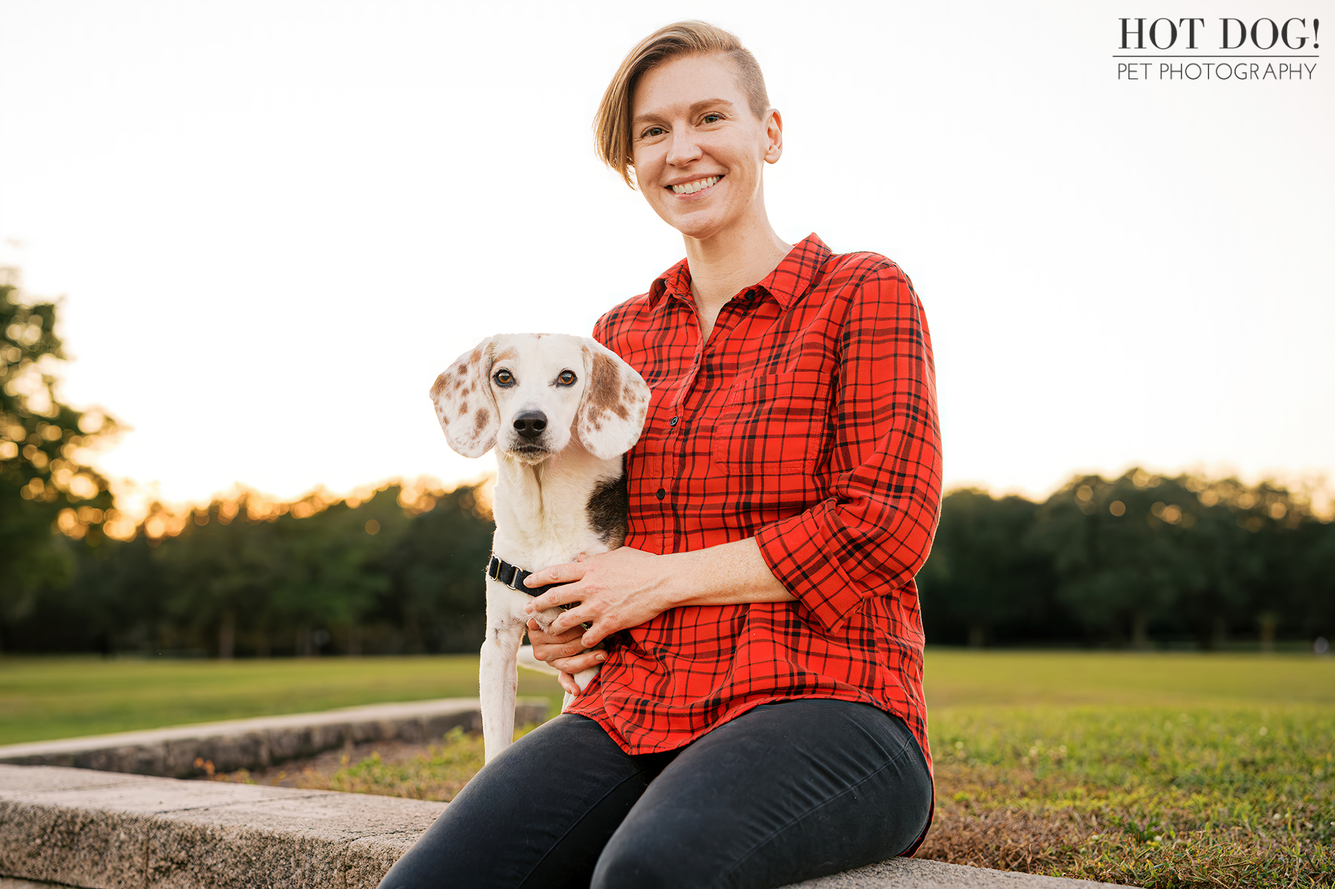 Smiling woman in a red plaid shirt sitting on a stone ledge while holding her senior beagle during a golden-hour pet session.