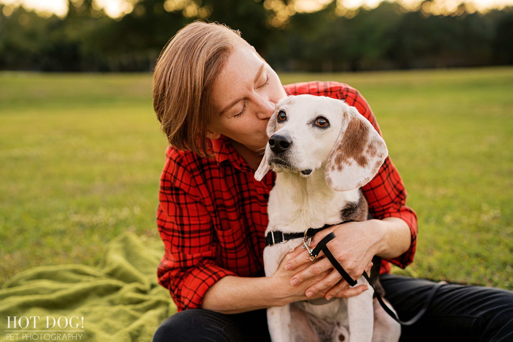 Woman kissing her senior beagle on the head while sitting together on a blanket outdoors, showing a close bond.