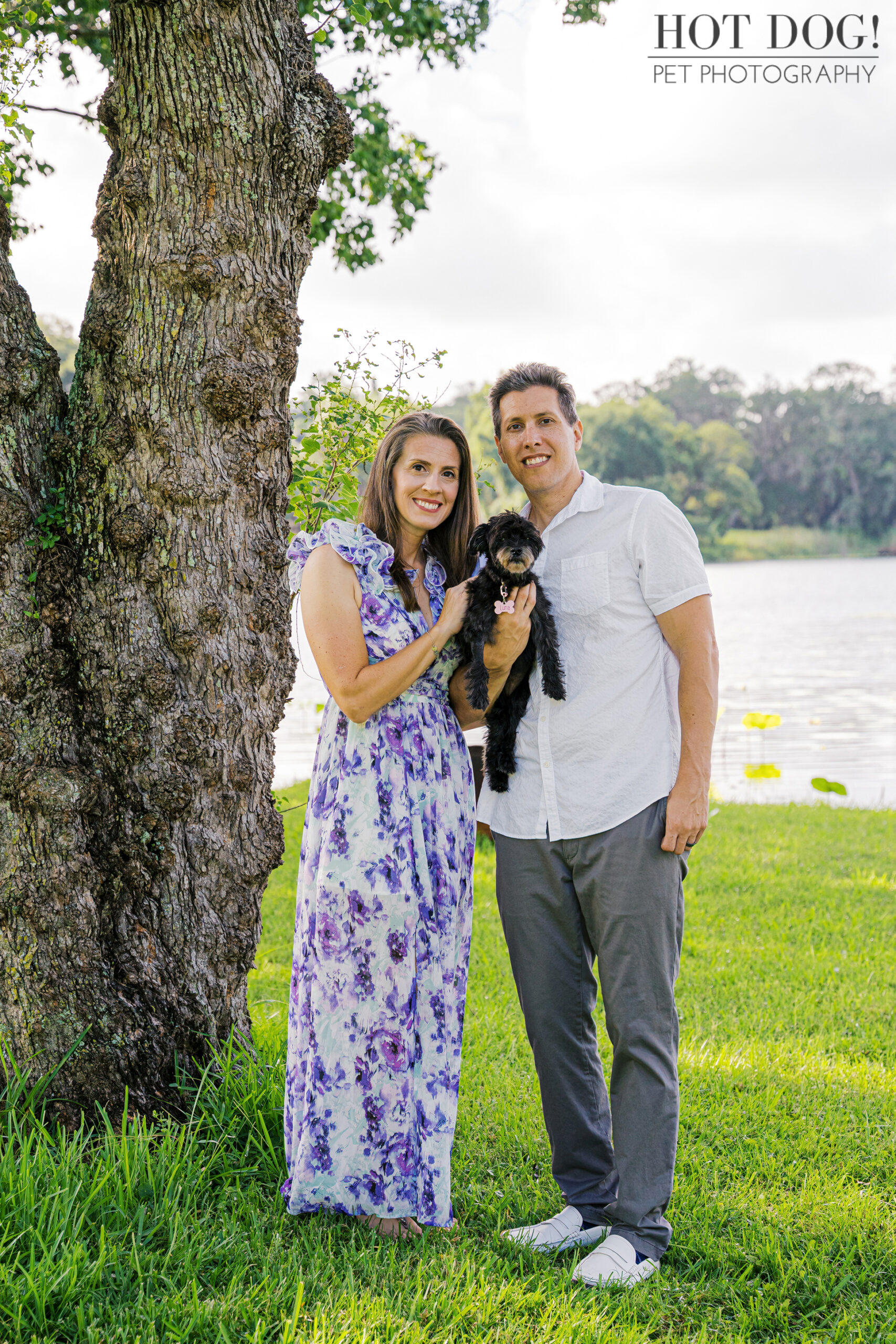 Family portrait by a tree with Rae and Nick holding Luna near a peaceful lake.