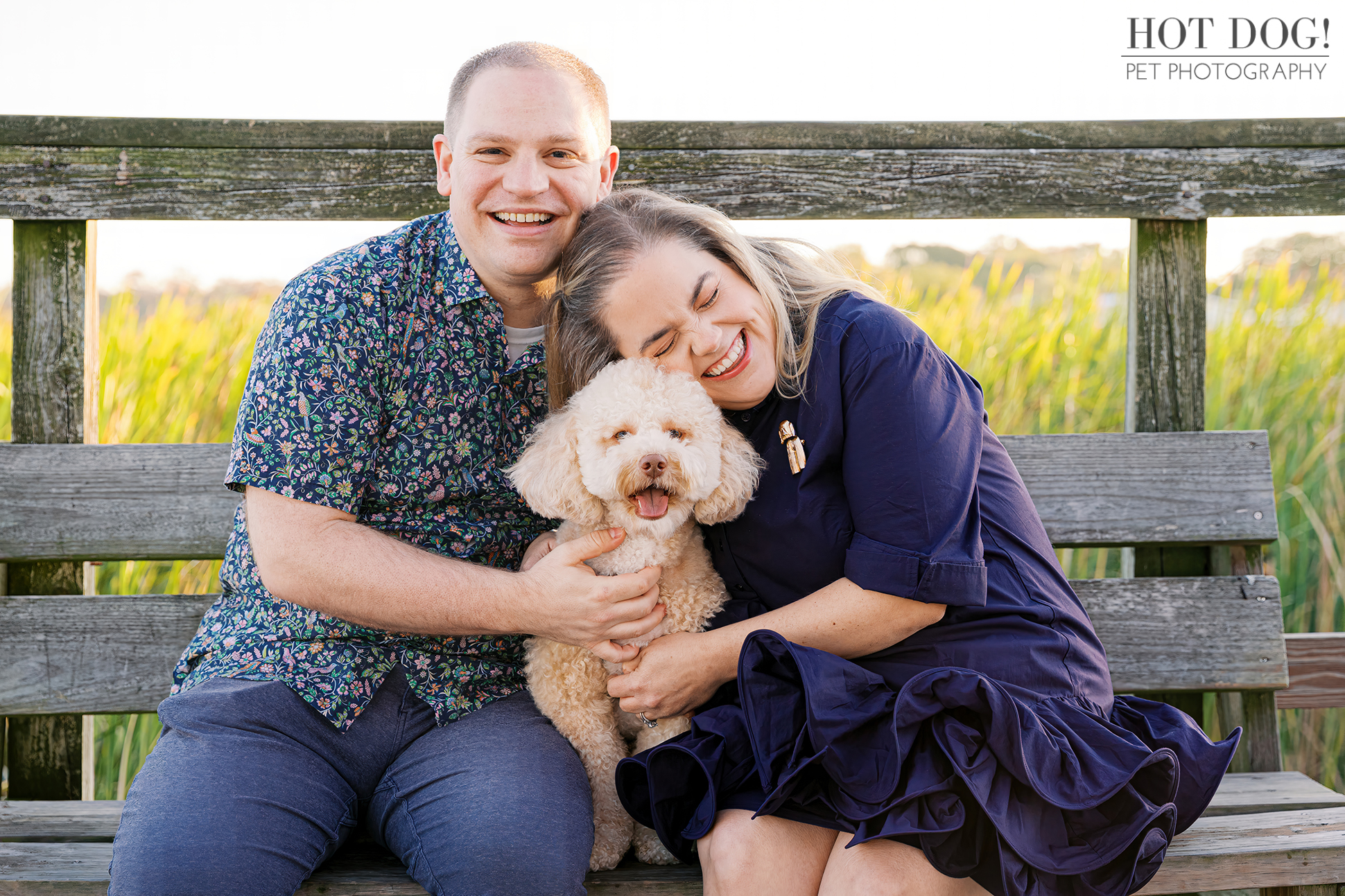 Lauren and Ian sit on a wooden dock bench at Newton Park with Mia between them, all smiling as golden light filters through tall grasses behind them.
