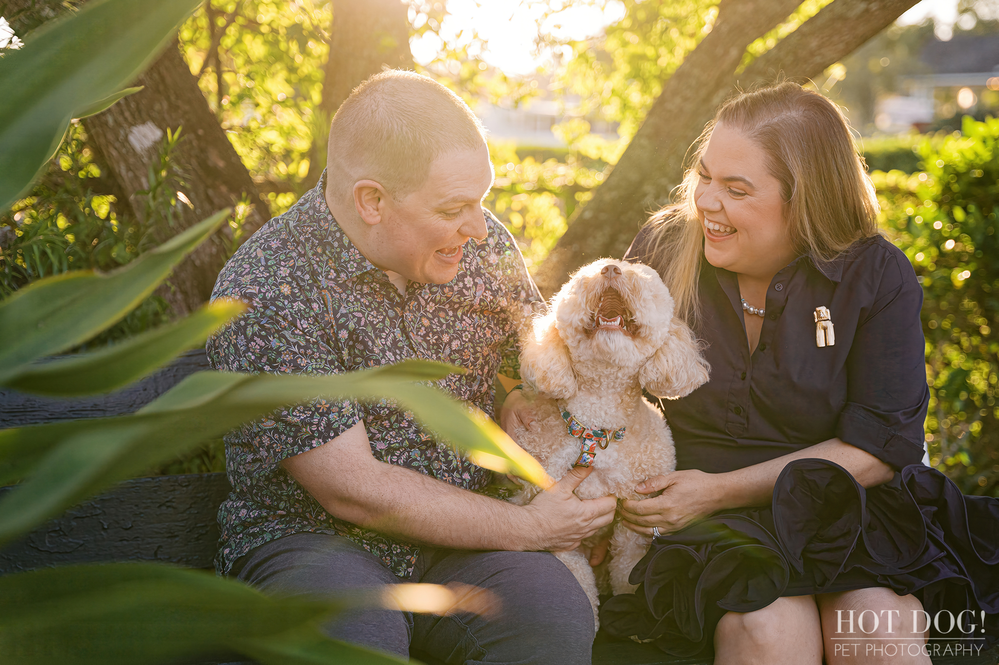 Lauren and Ian laugh together on a shaded bench while Mia looks up at them with a big smile, sunlight streaming through leaves around the trio.