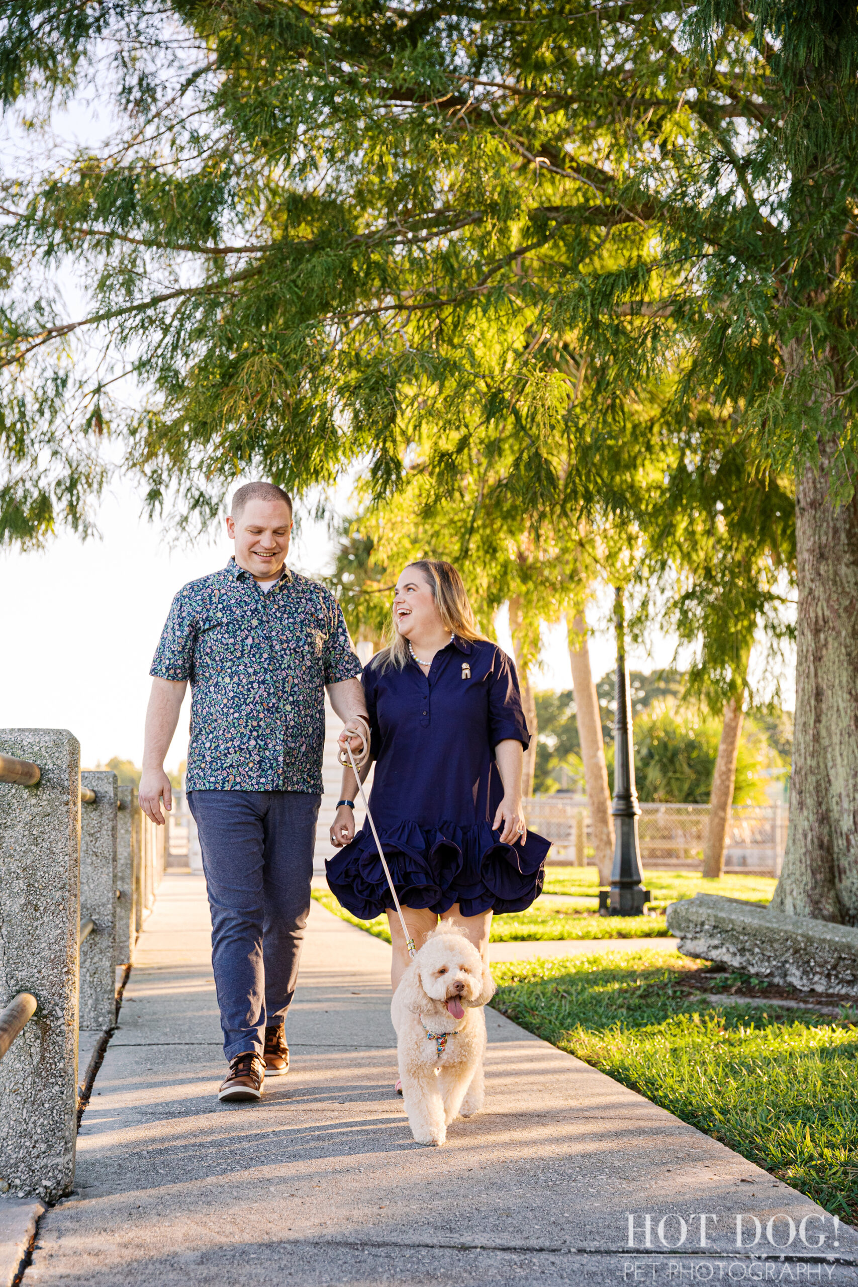 Lauren and Ian walk hand-in-hand along the lakeside path at Newton Park while Mia trots ahead on her leash, the trees glowing with warm morning light.
