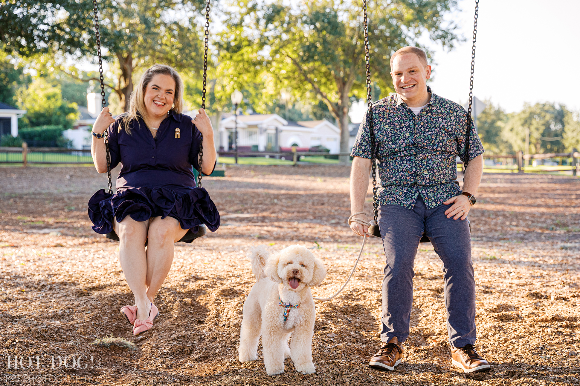 Lauren and Ian sit on adjacent playground swings, smiling toward the camera, while Mia stands between them on the wood chips with her tongue out.