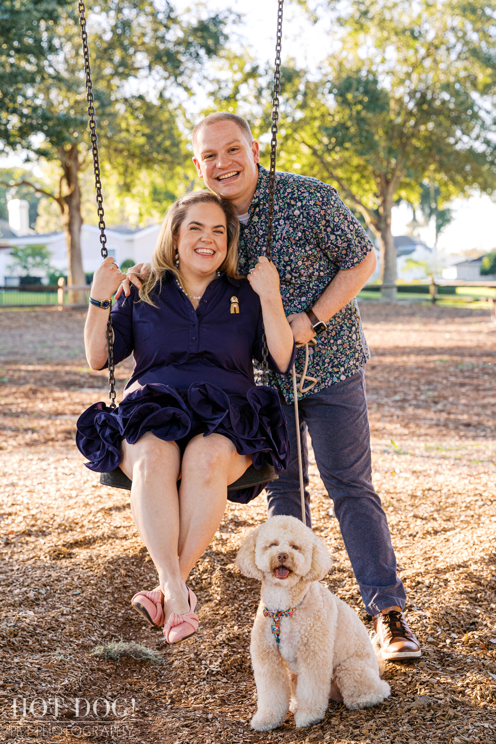 Lauren sits on a playground swing smiling while Ian stands behind her, holding the swing chain, as Mia sits happily at their feet on the wood chips.
