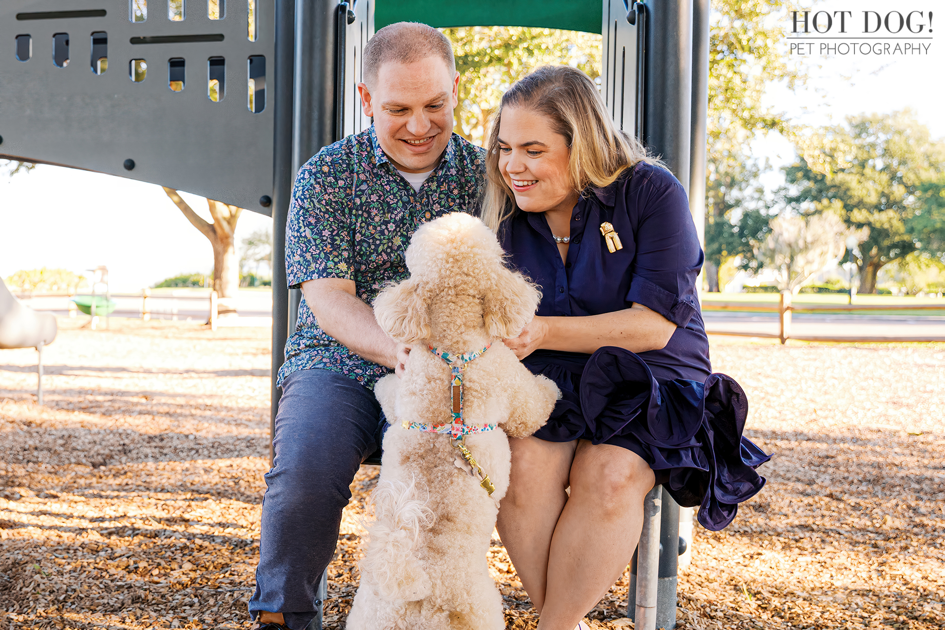 Lauren and Ian sit at the bottom of the playground structure, laughing as Mia stands on her hind legs with her front paws on their knees.