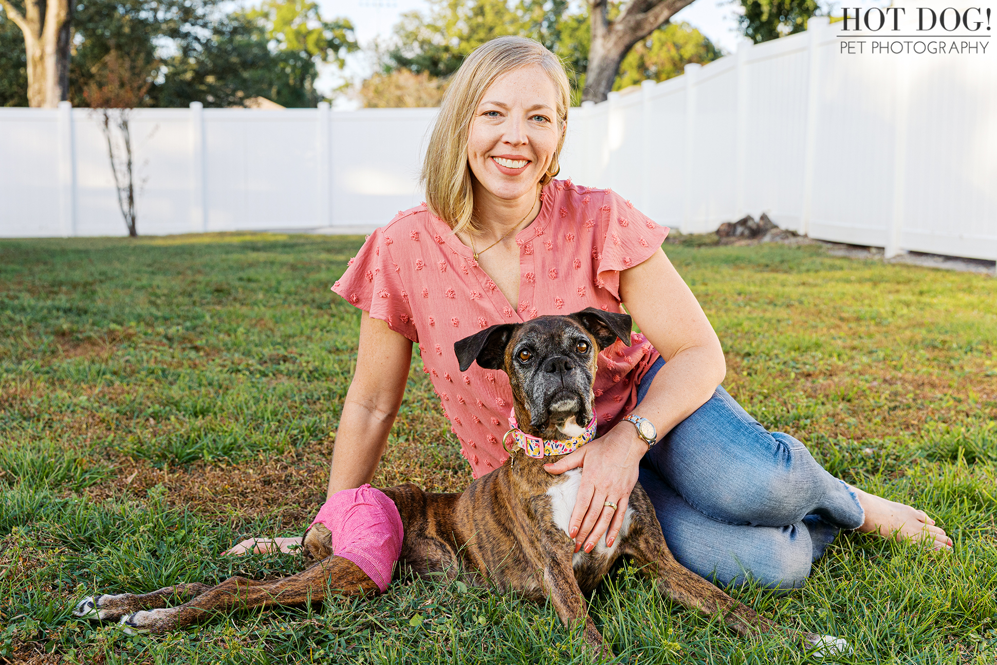 Woman sitting on the grass beside her senior Boxer in a fenced backyard.