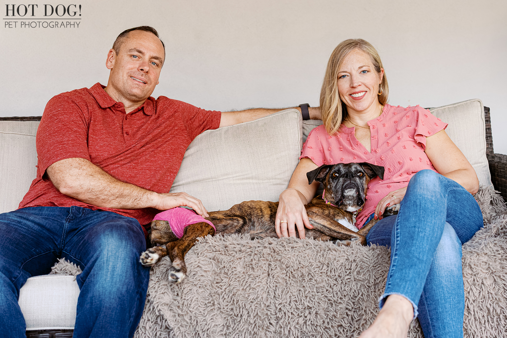 Parents sitting on a couch indoors with their senior Boxer resting between them.