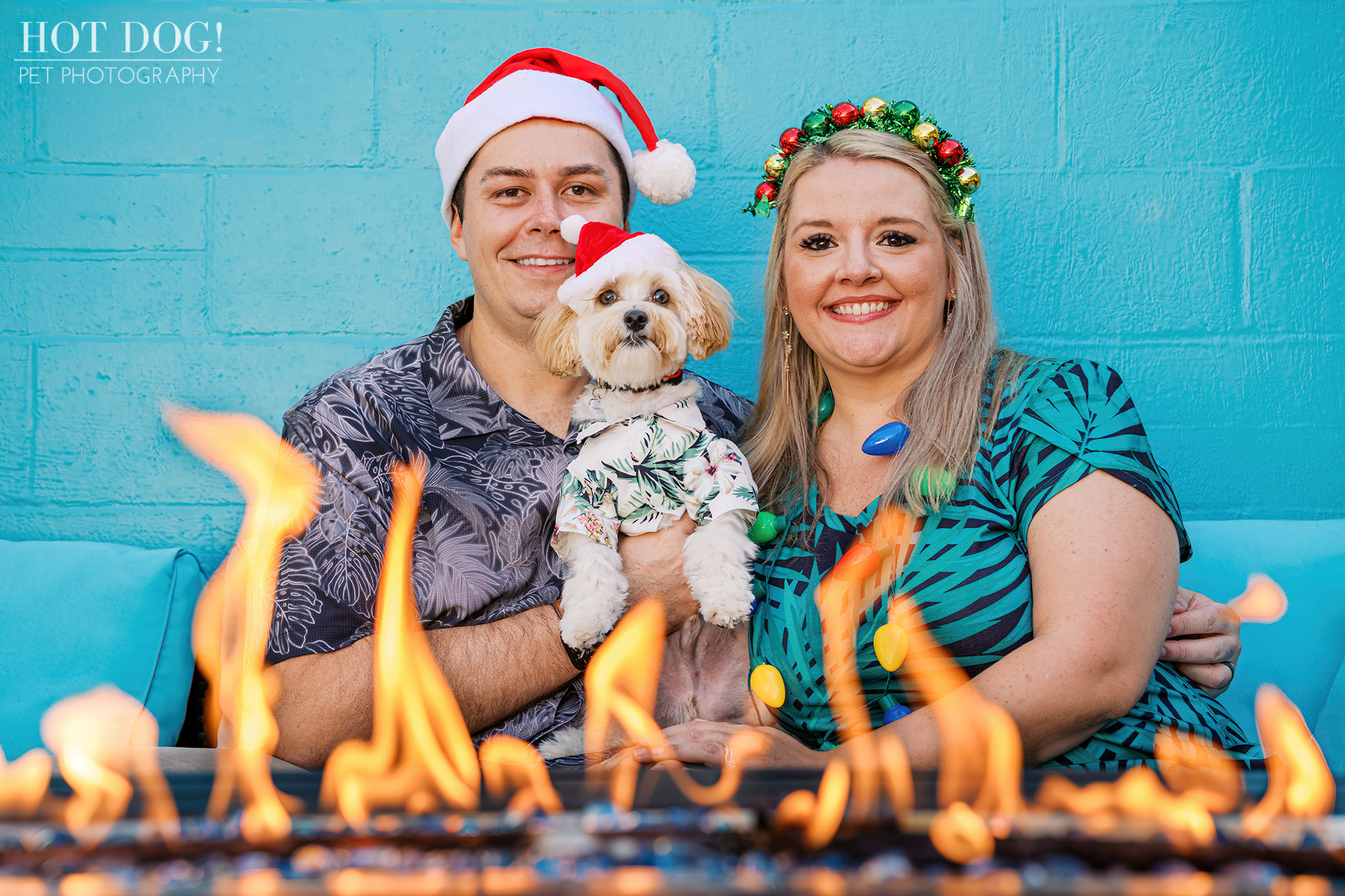 Julie and Remi sitting by an outdoor fire feature with Milo in a Santa hat, all smiling in front of a bright blue wall.