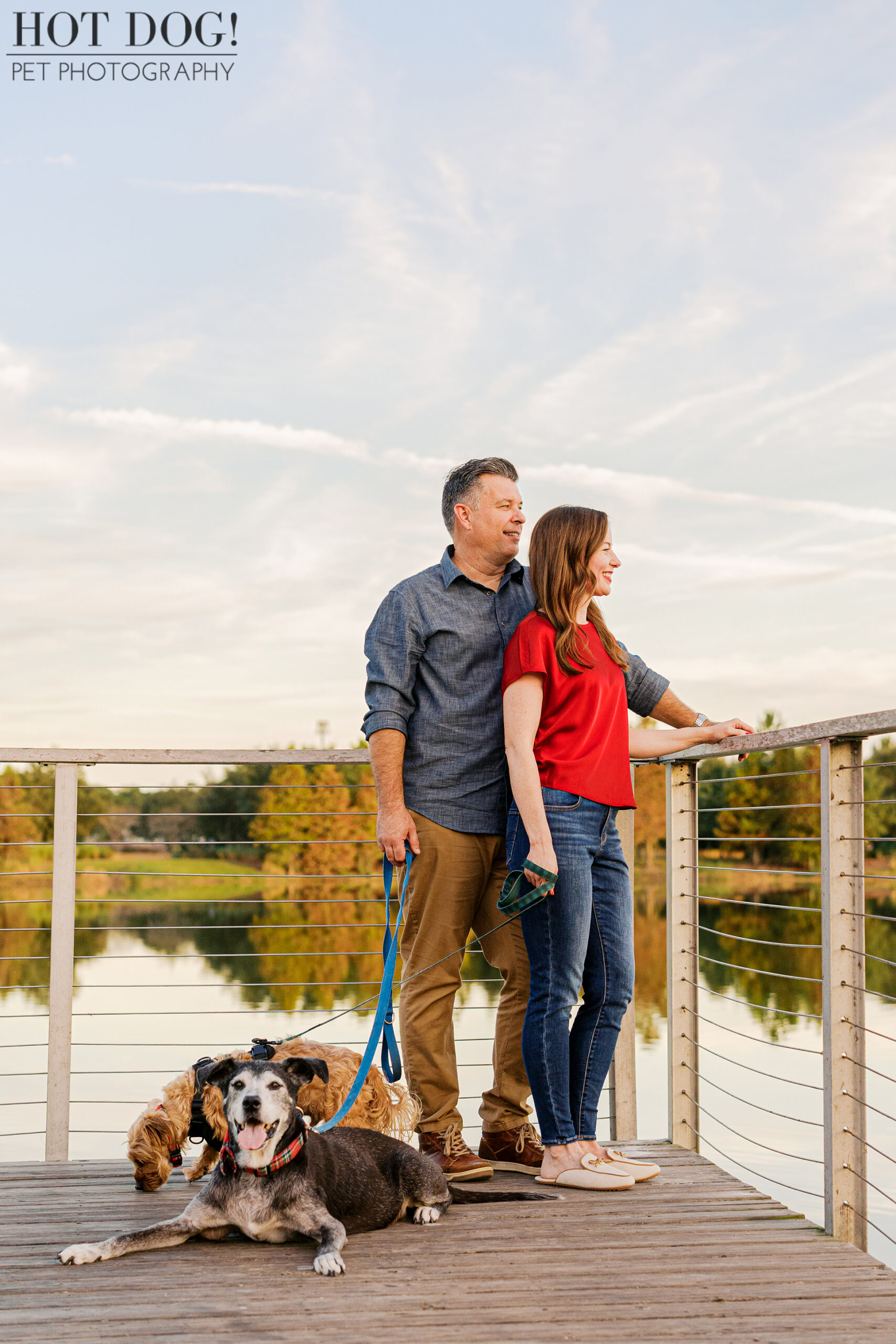 Couple standing together on a wooden bridge holding dog leashes, with their two dogs resting at their feet near the water.
