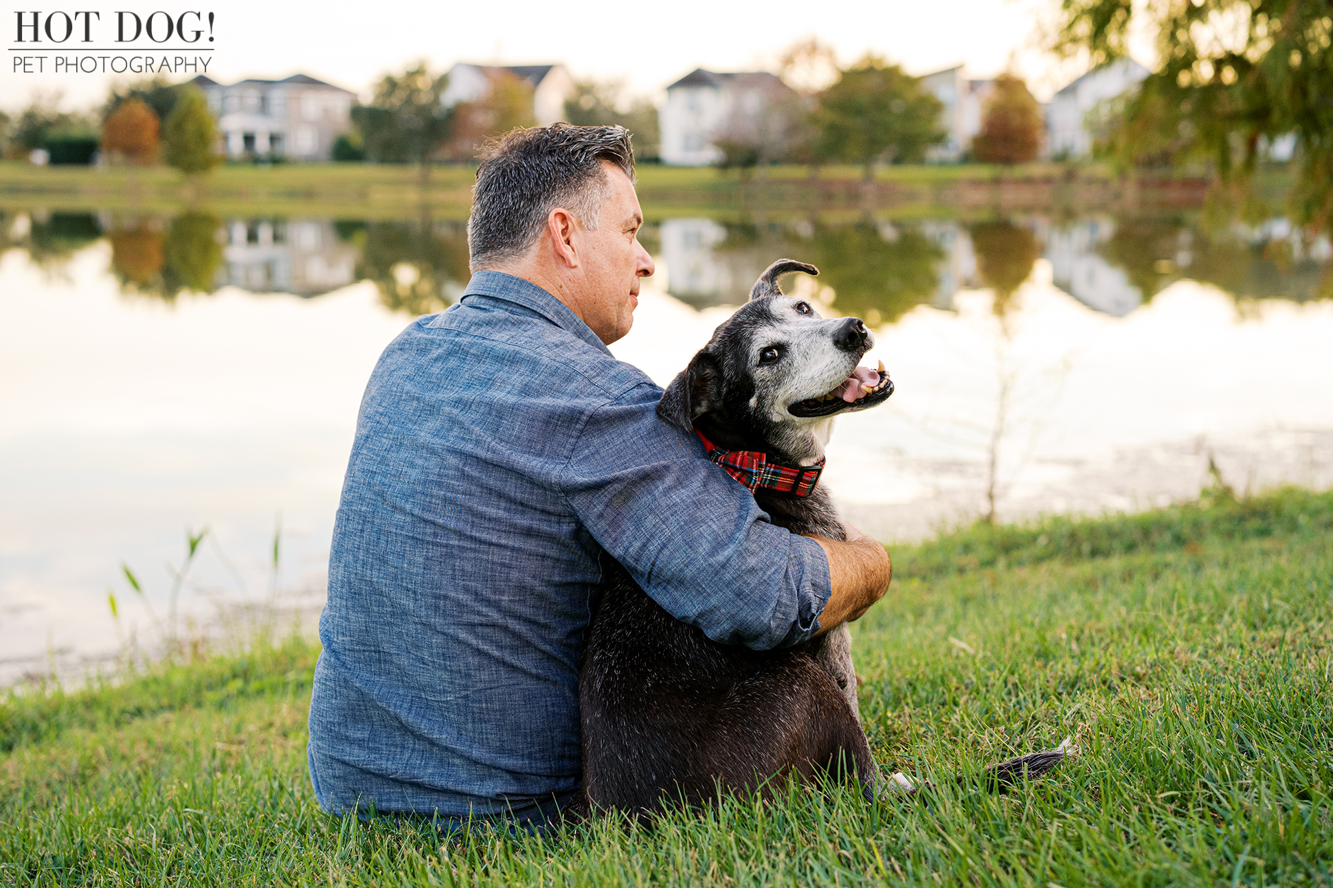 Adult man sitting on grass by a lakeside holding his senior black and white lab mix close, photographed during a calm outdoor pet photography session.