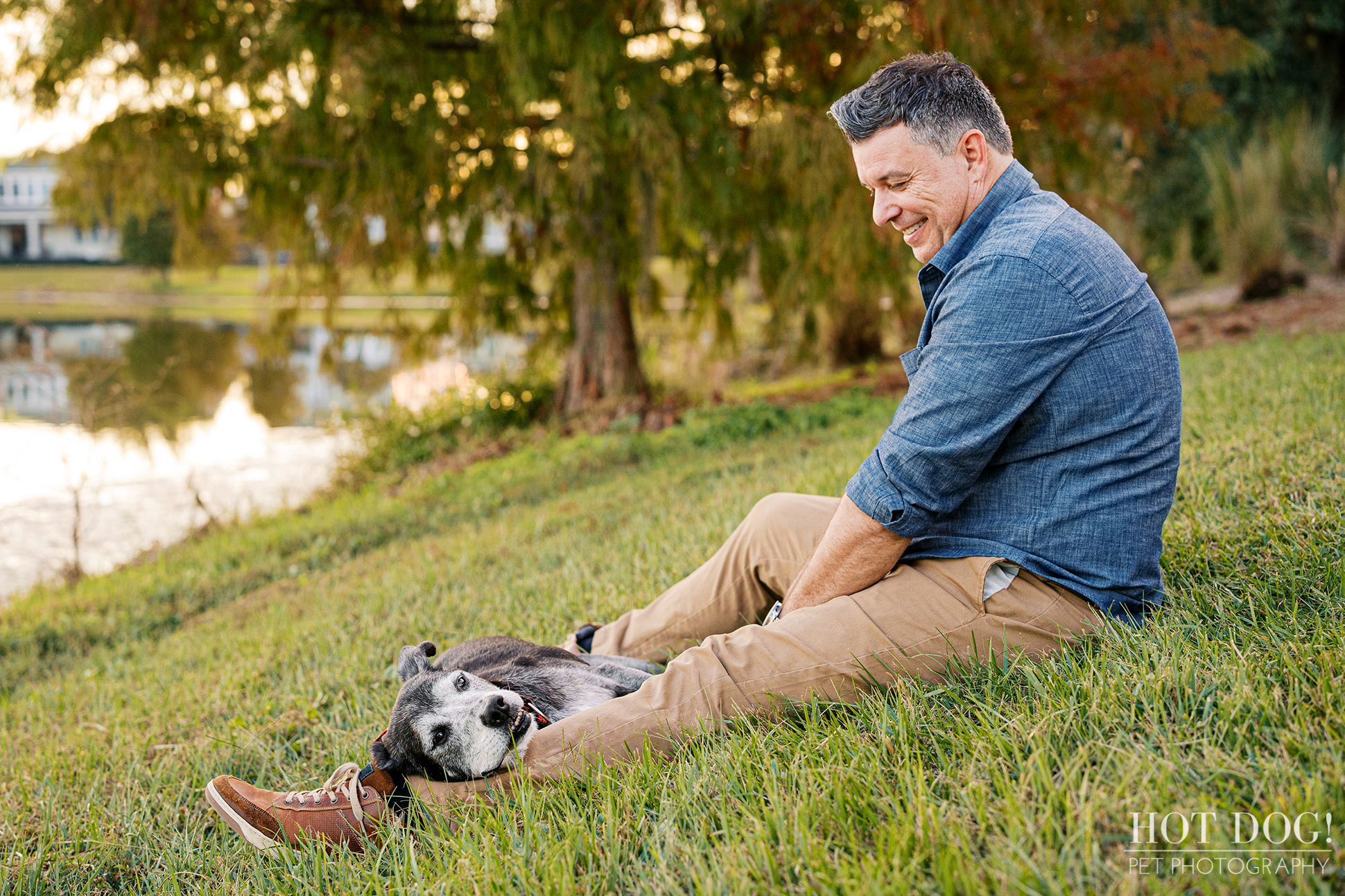 Adult man sitting on a grassy slope smiling down at his senior black and white lab mix resting near his legs, with a lake and trees in the background.