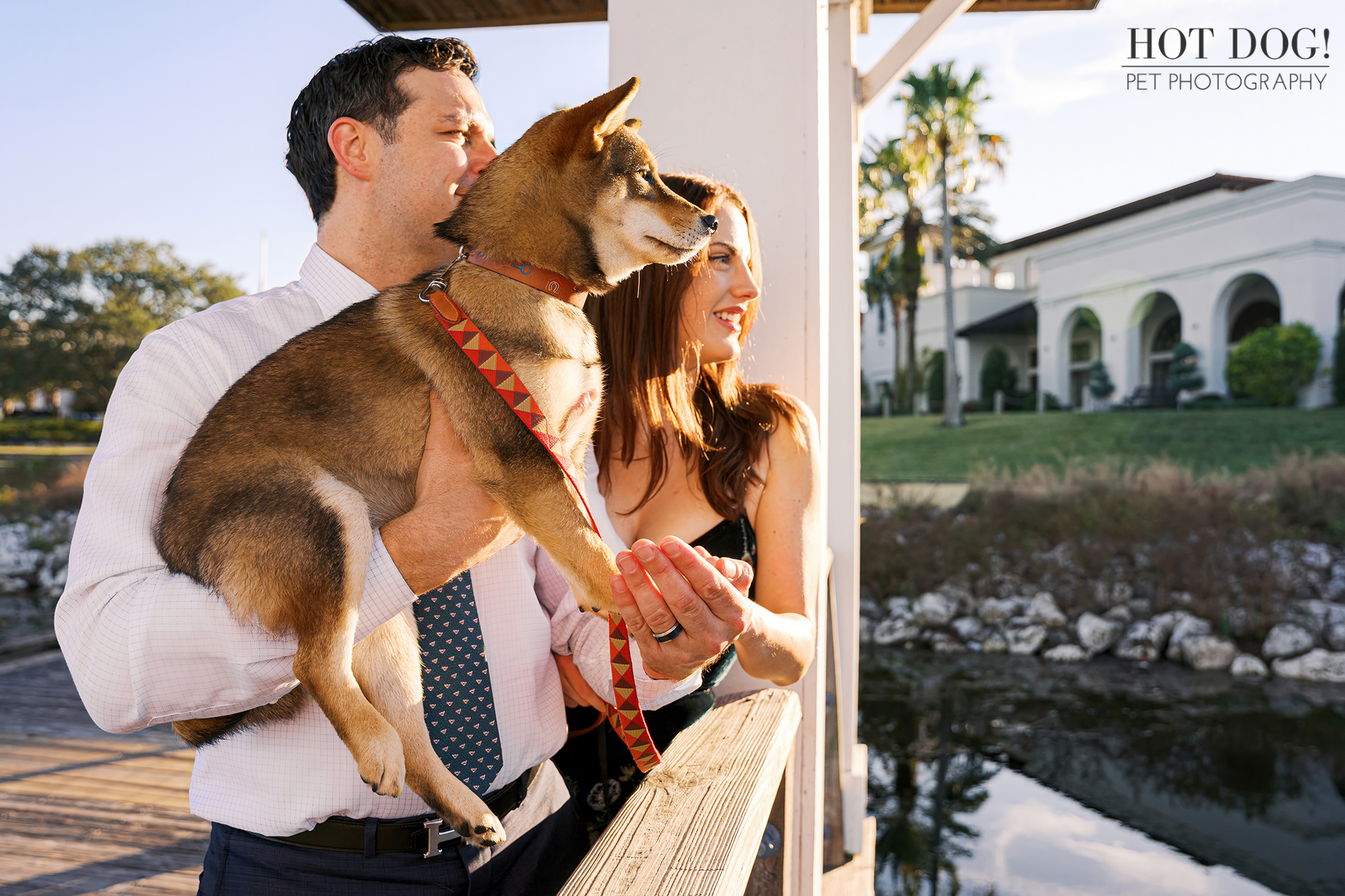 Man holding a sesame Shiba Inu puppy while standing beside a woman on a wooden boardwalk near water during an outdoor pet photography session.