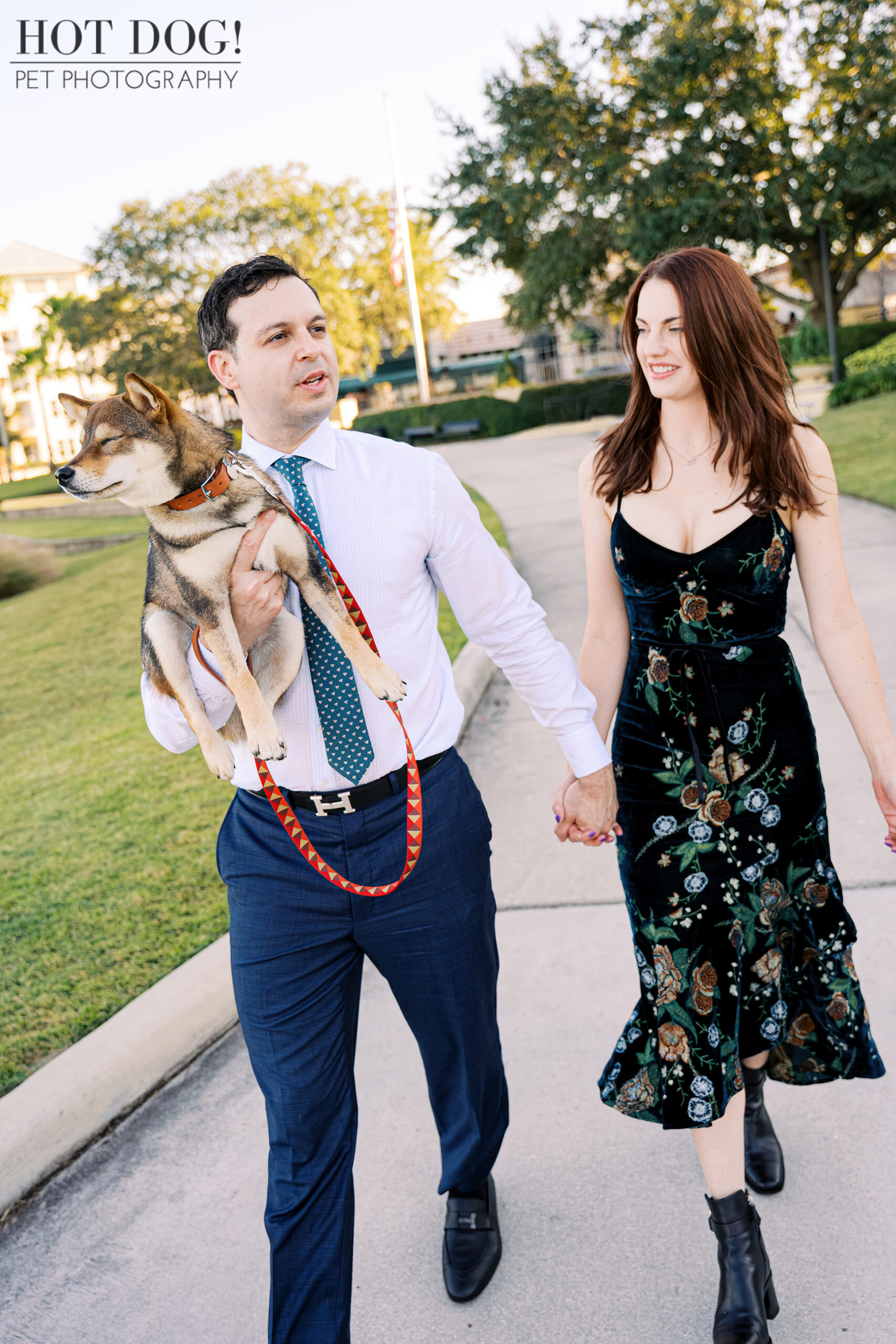 Couple walking hand in hand along a park path while one carries their sesame Shiba Inu puppy during an outdoor photo session.
