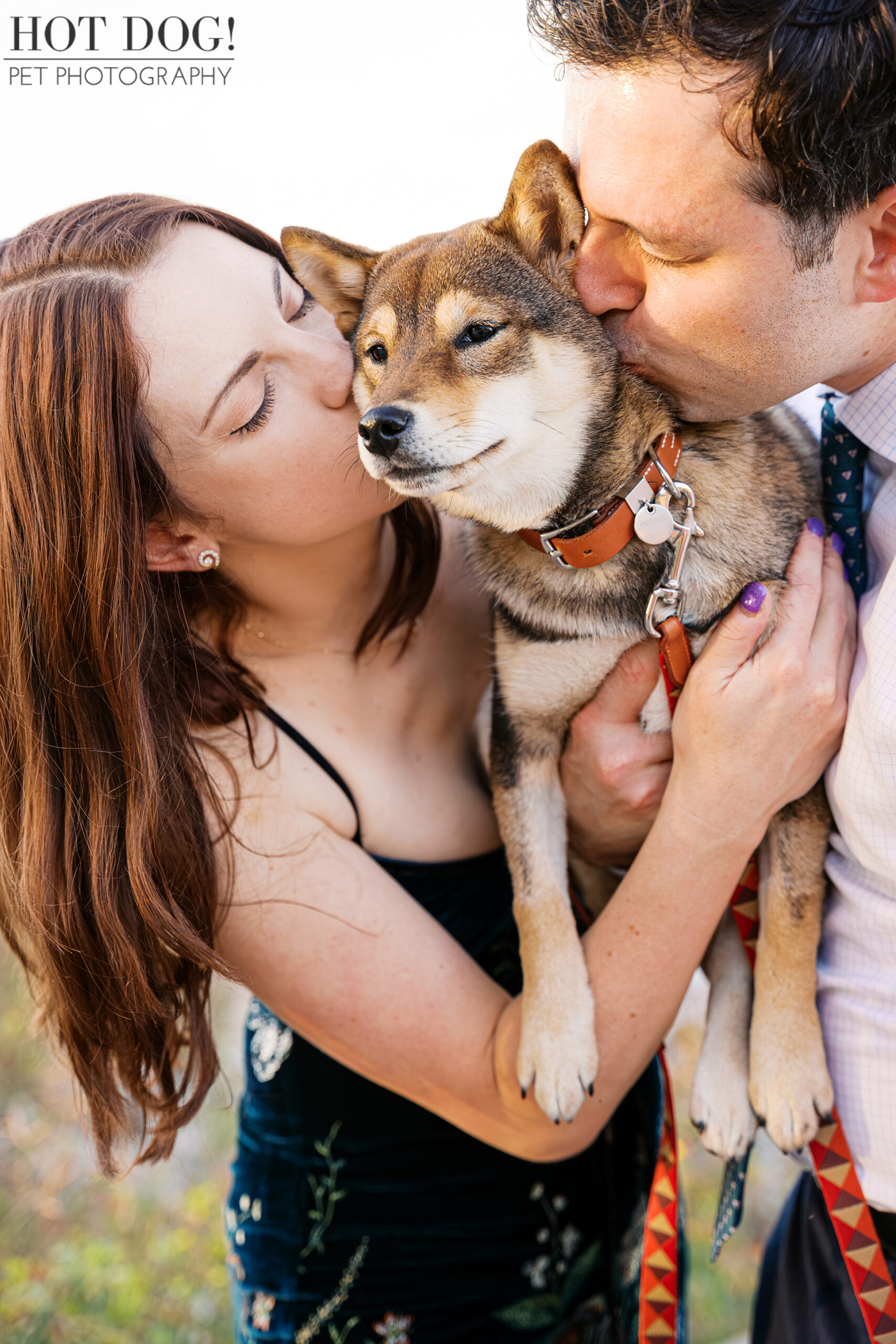 Close-up of a sesame Shiba Inu puppy being held while both owners kiss her cheeks during an outdoor pet photography session.