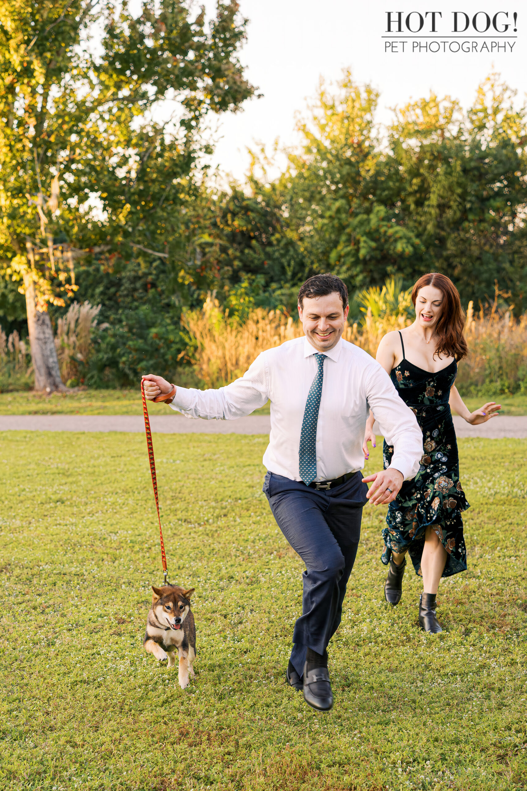 Man running playfully across grass while holding a leash, followed by a woman, as their sesame Shiba Inu puppy trots ahead during a pet photography session.