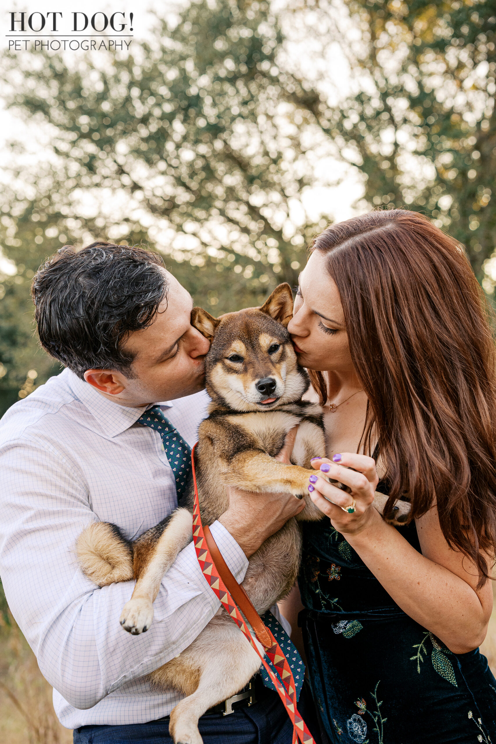 Close-up of a sesame Shiba Inu puppy being held as both owners gently kiss her cheeks, photographed outdoors with soft greenery behind.