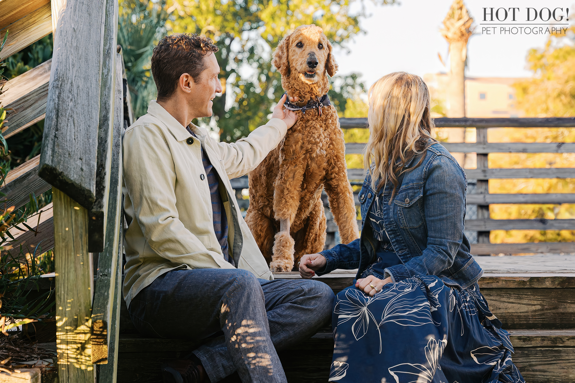 Jen and Cory sitting on wooden steps at Gaston Edwards Park with Wizard, their apricot Goldendoodle, standing between them while Cory gently pets him.
