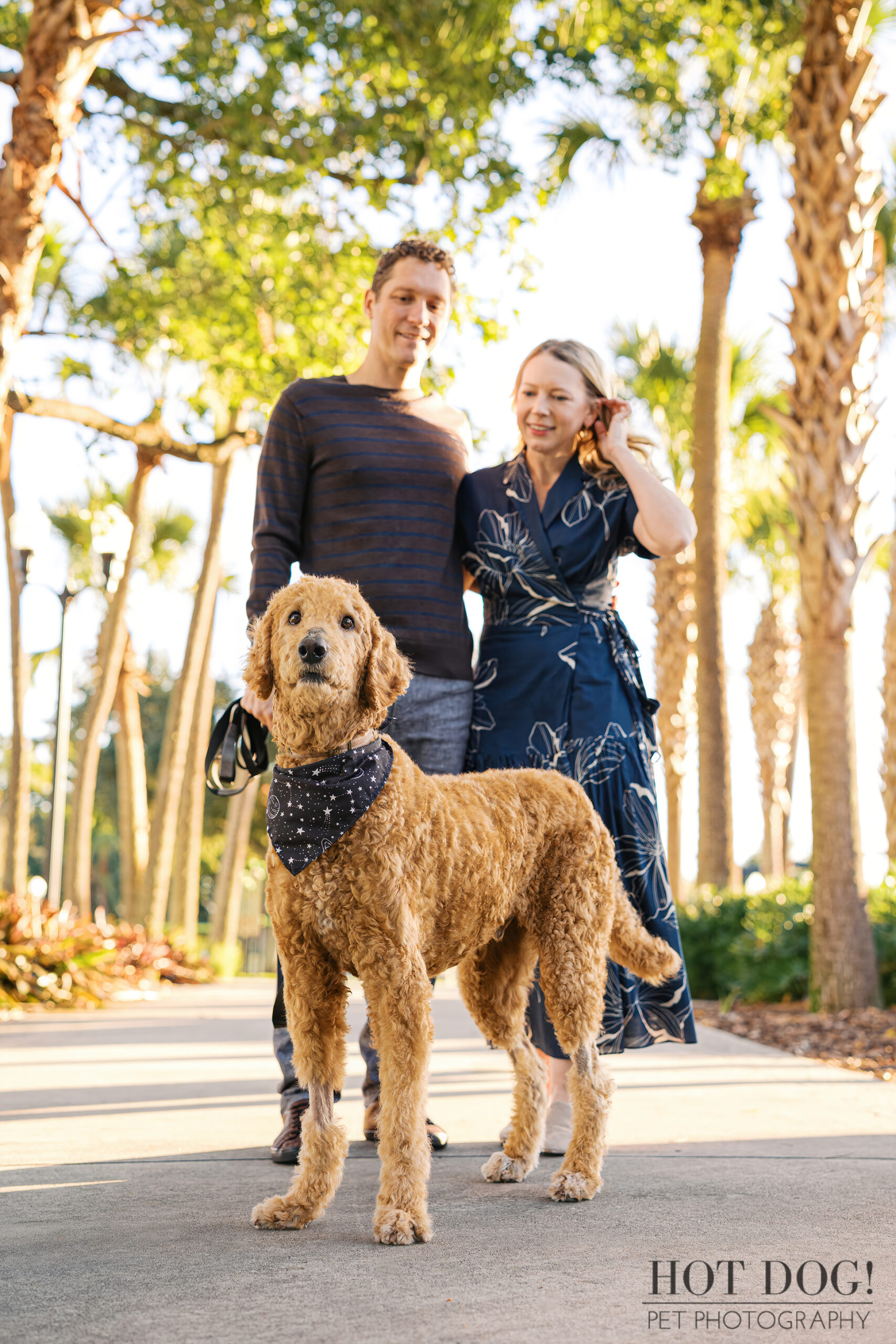 Wizard standing proudly in front of Jen and Cory on the lakeside walkway, wearing a navy star-patterned bandana as the couple smiles behind him.