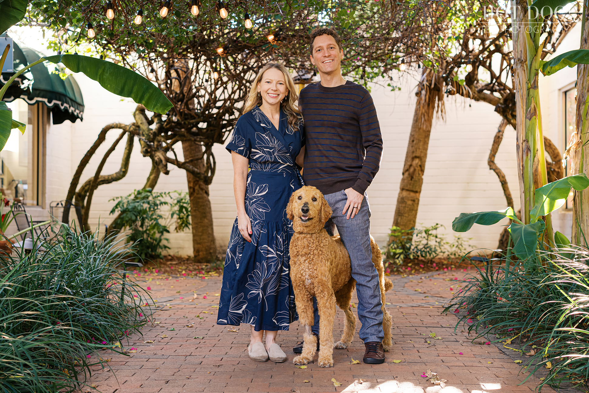 Jen and Cory standing together in a brick courtyard with string lights overhead, smiling as Wizard stands proudly between them.