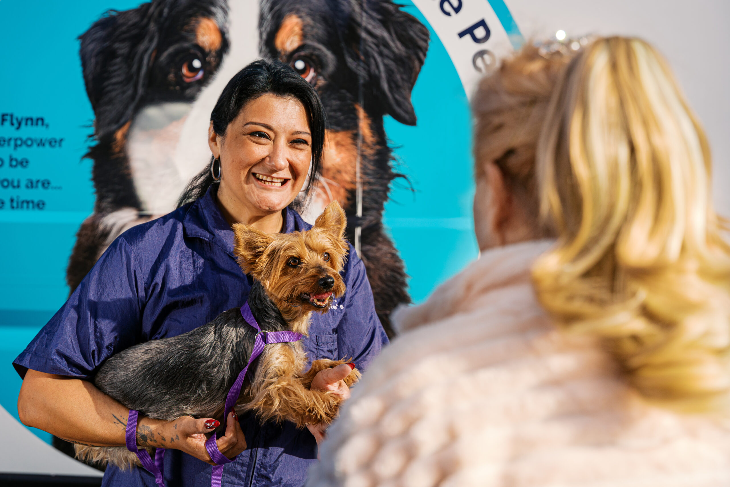 Mobile pet groomer holding a Yorkshire Terrier while speaking with a client in front of a branded Woofie’s grooming van during a commercial pet photography session.
